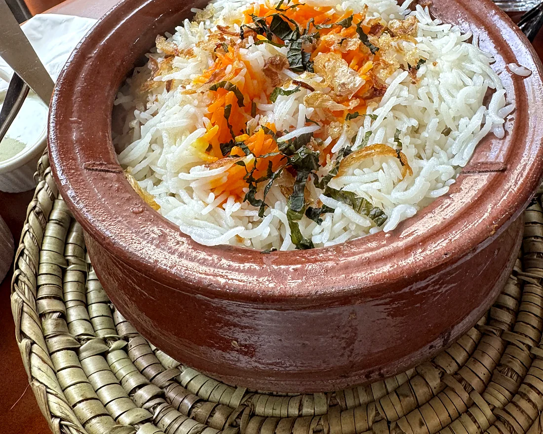 Clay pot of white basmati biryani topped with saffron rice and fried onions, on a woven mat