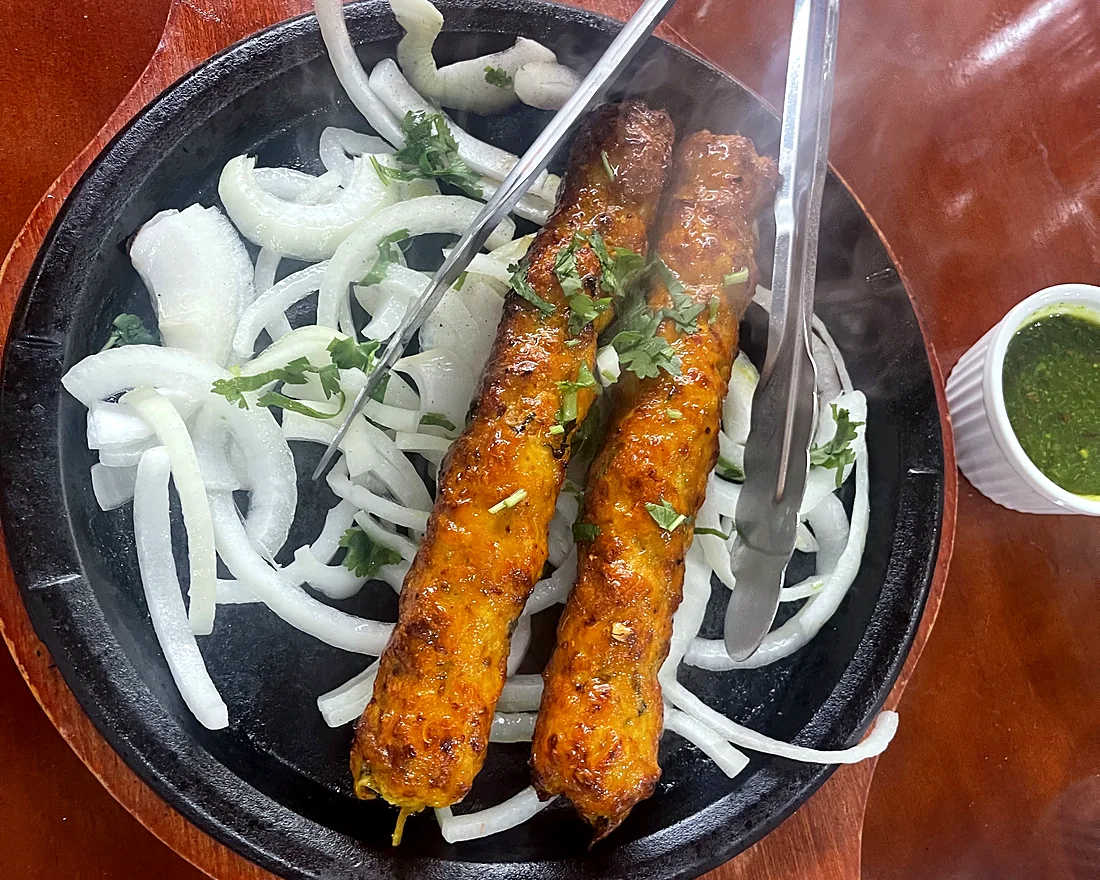 Two chicken seekh kababs on a sizzling iron plate with white onion rings, tongs and green chutney