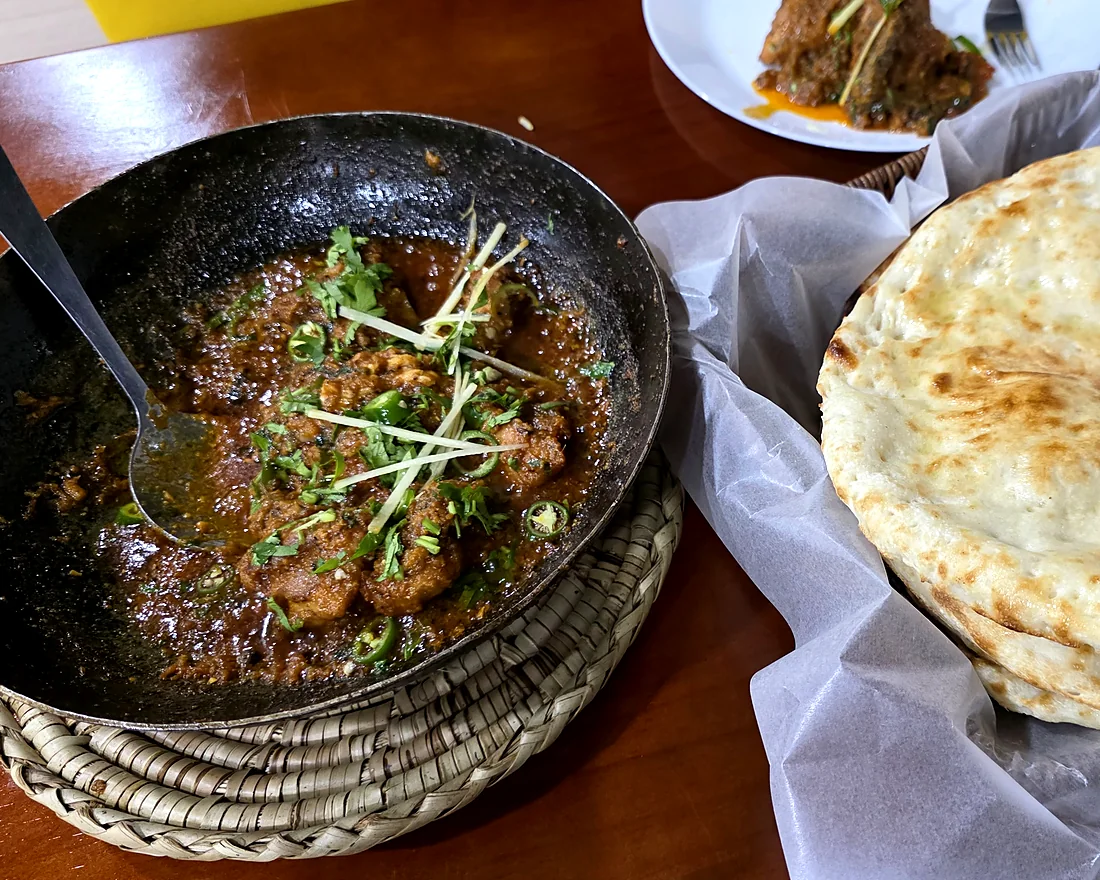 Karahi in a black iron wok on a woven mat next to naan in a paper-lined basket