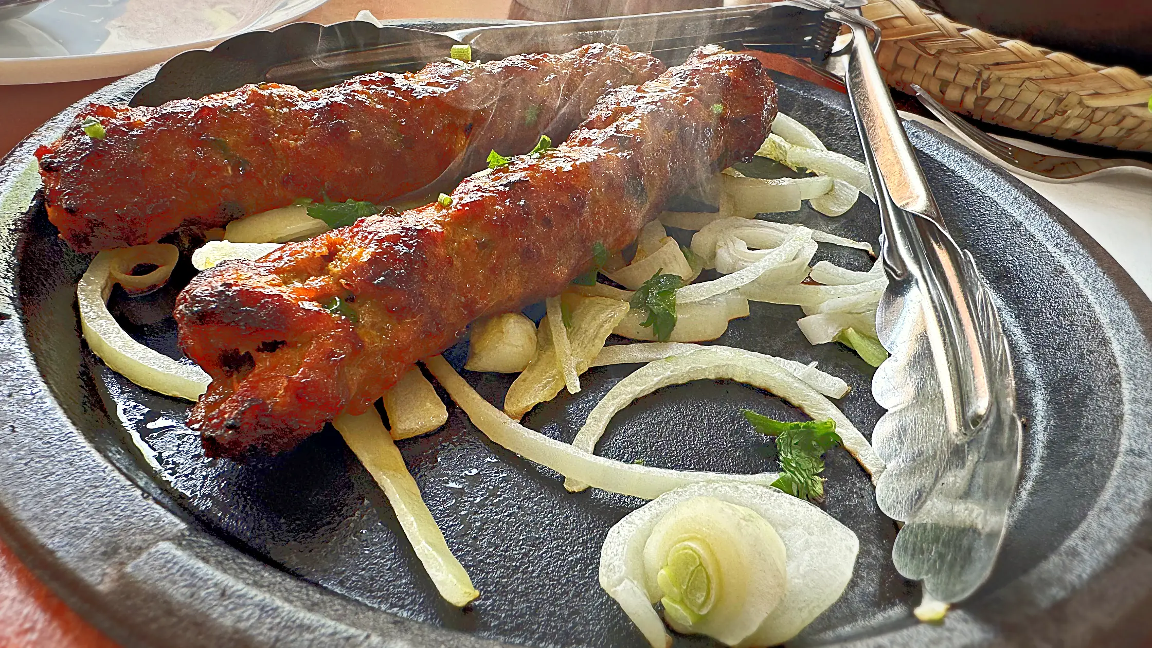 Two seekh kababs on a sizzling iron plate with onion rings and serving tongs
